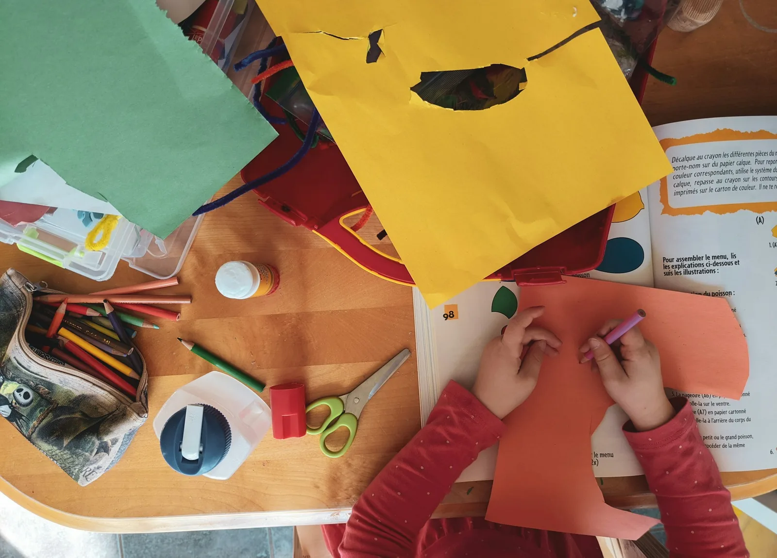 Overhead view of a table with colored paper, scissors and pencils while a child cuts something out