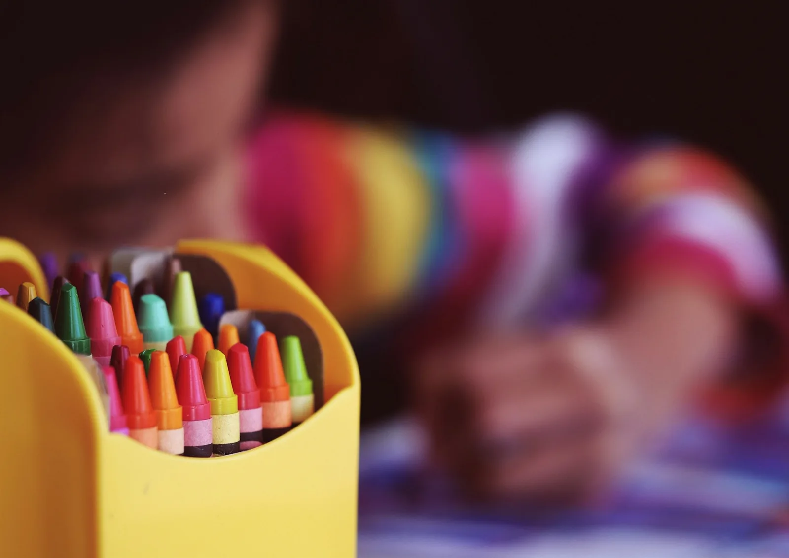 Yellow box of colored crayons in the foreground with a child drawing behind